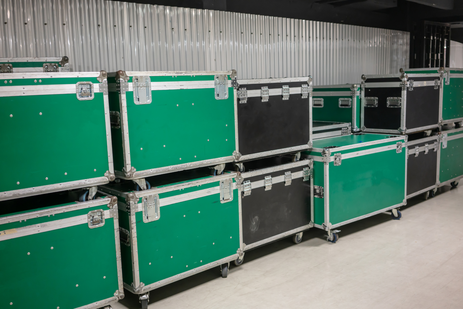 Green and black fight cases stack up in front of a corrugated metal partition in two organized rows.