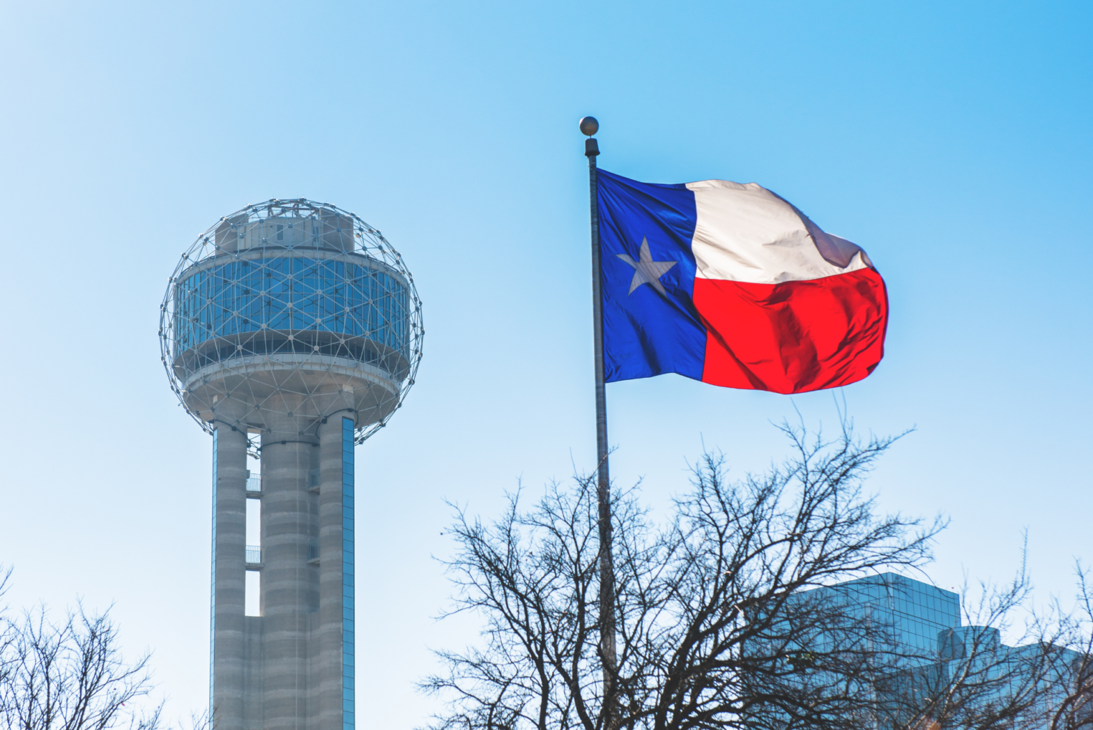 A Texas flag blows in the wind next to Dallas’s Reunion Tower in the background on a bright, sunny day.